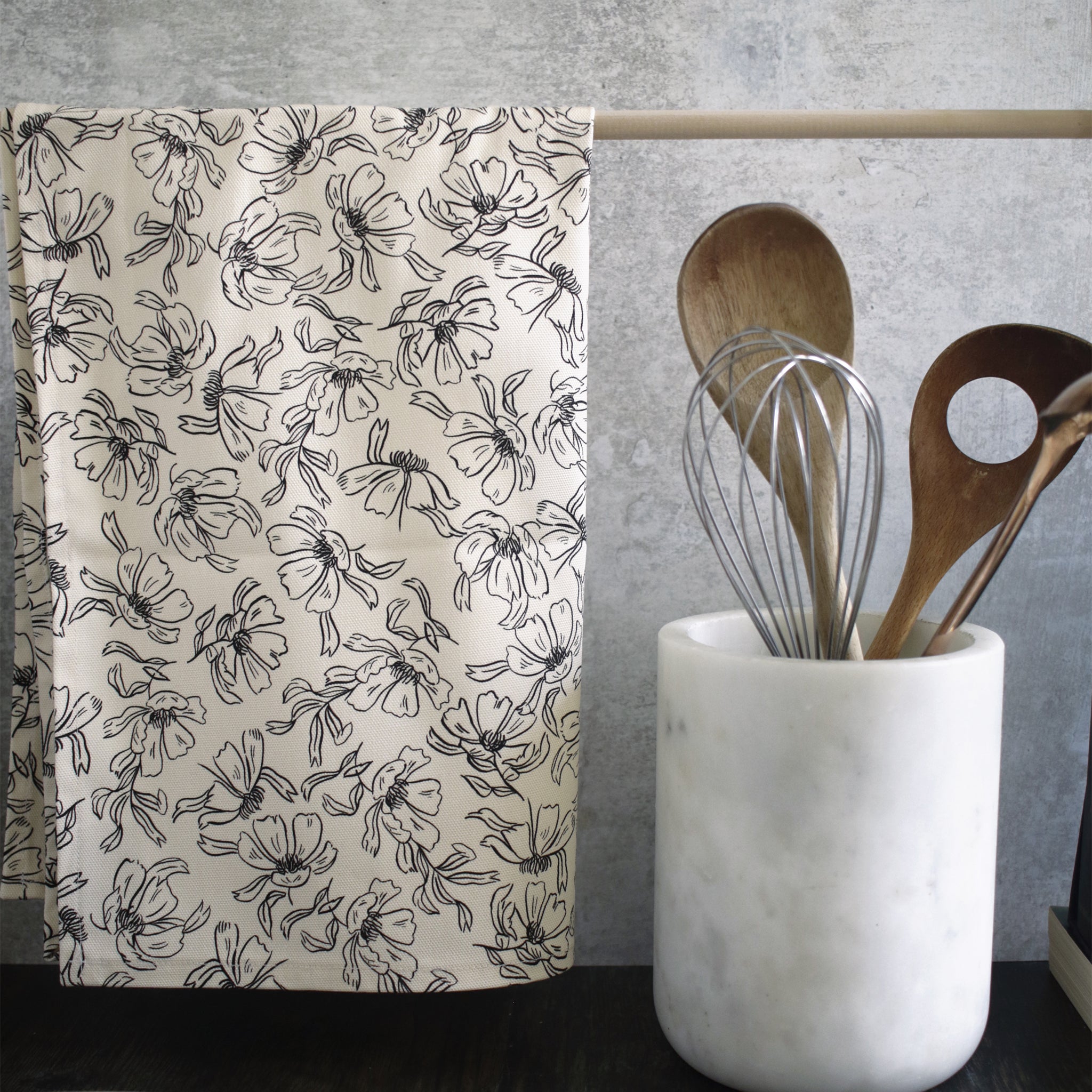 A black and cream floral patterned tea towel, hanging from a wooden rail on a countertop