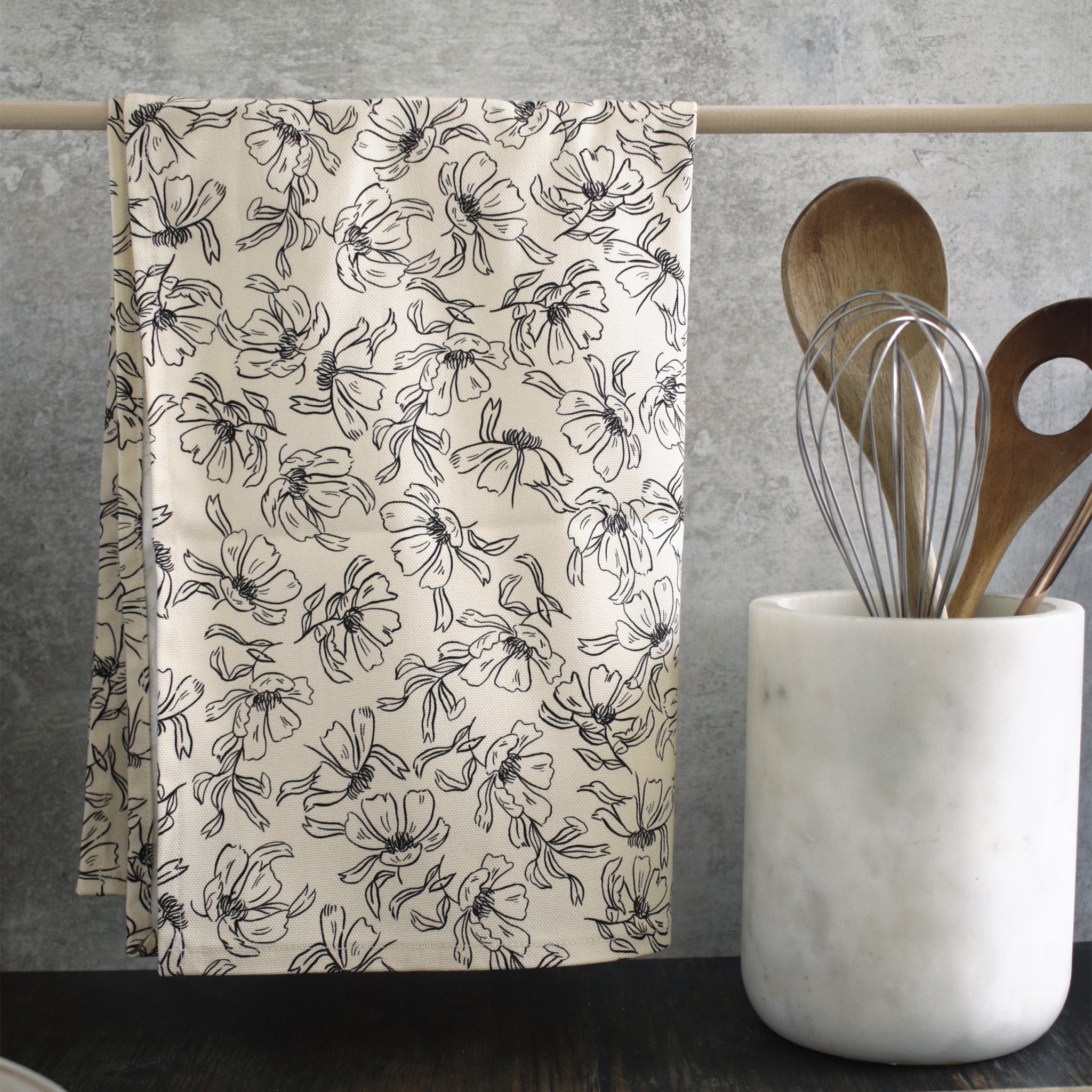 A black and cream floral patterned tea towel, hanging from a wooden rail on a countertop