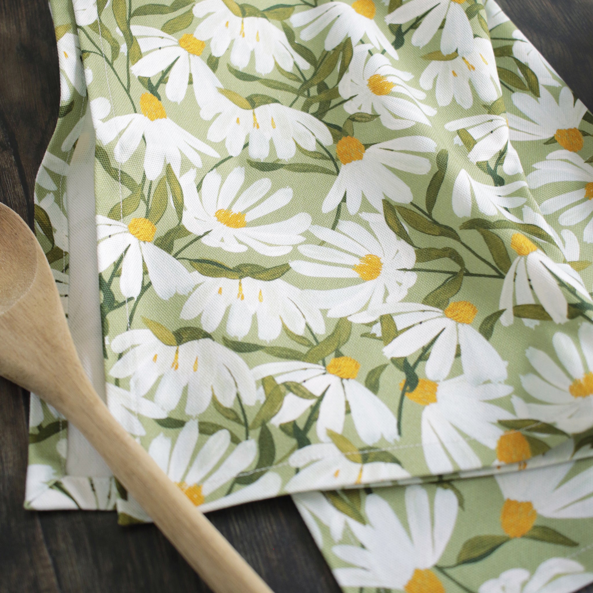a green cotton tea towel, covered in painted white daisy flowers, sitting on a dark wood table alongside plates and utensils