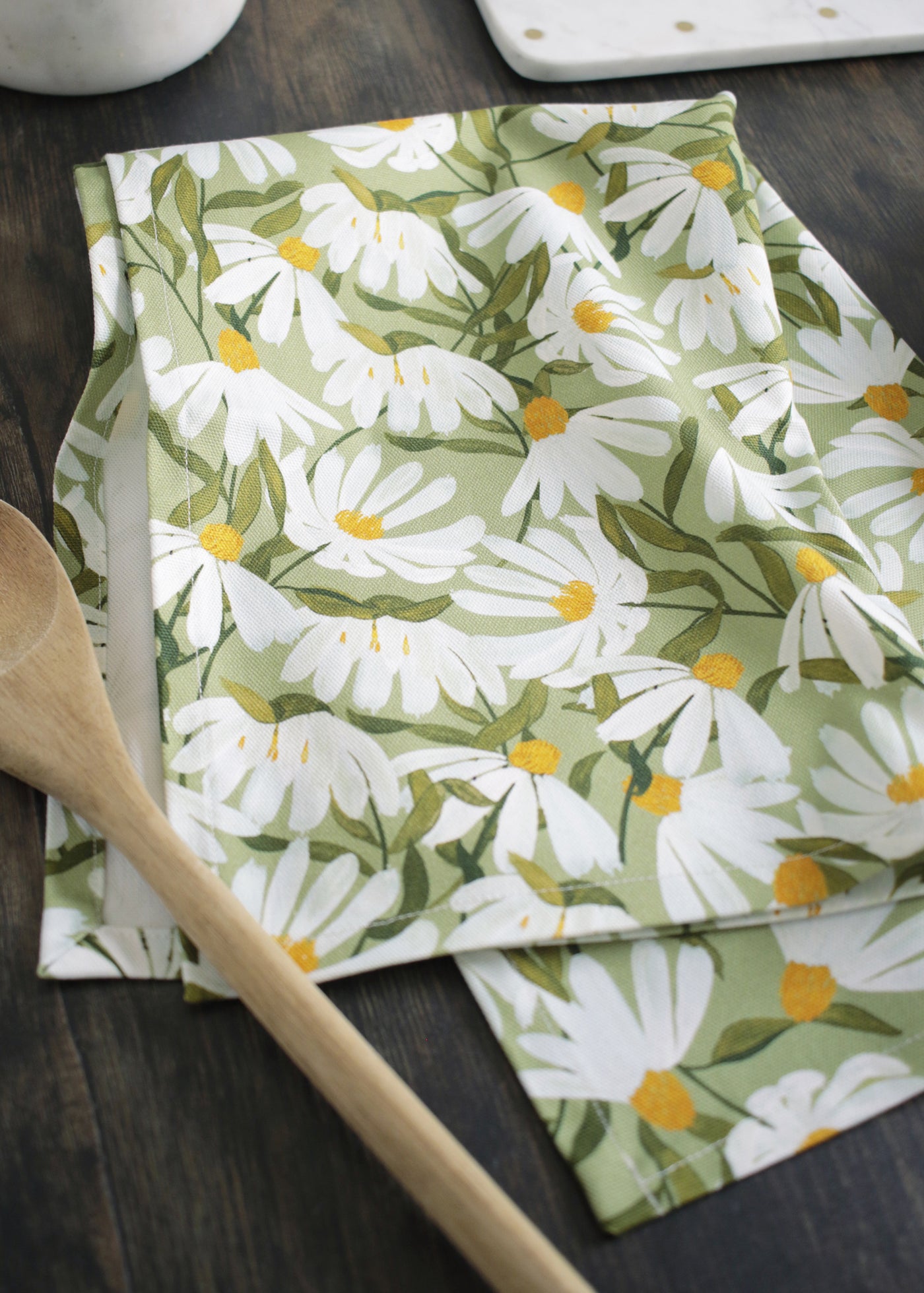a green cotton tea towel, covered in painted white daisy flowers, sitting on a dark wood table alongside plates and utensils