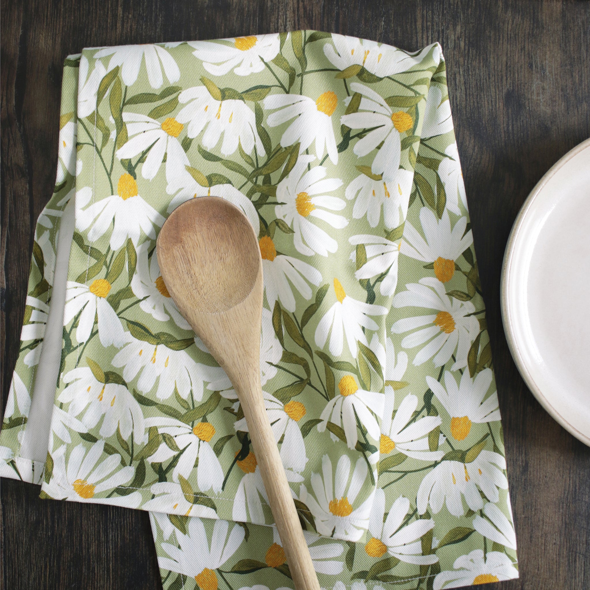 a green cotton tea towel, covered in painted white daisy flowers, sitting on a dark wood table alongside plates and utensils