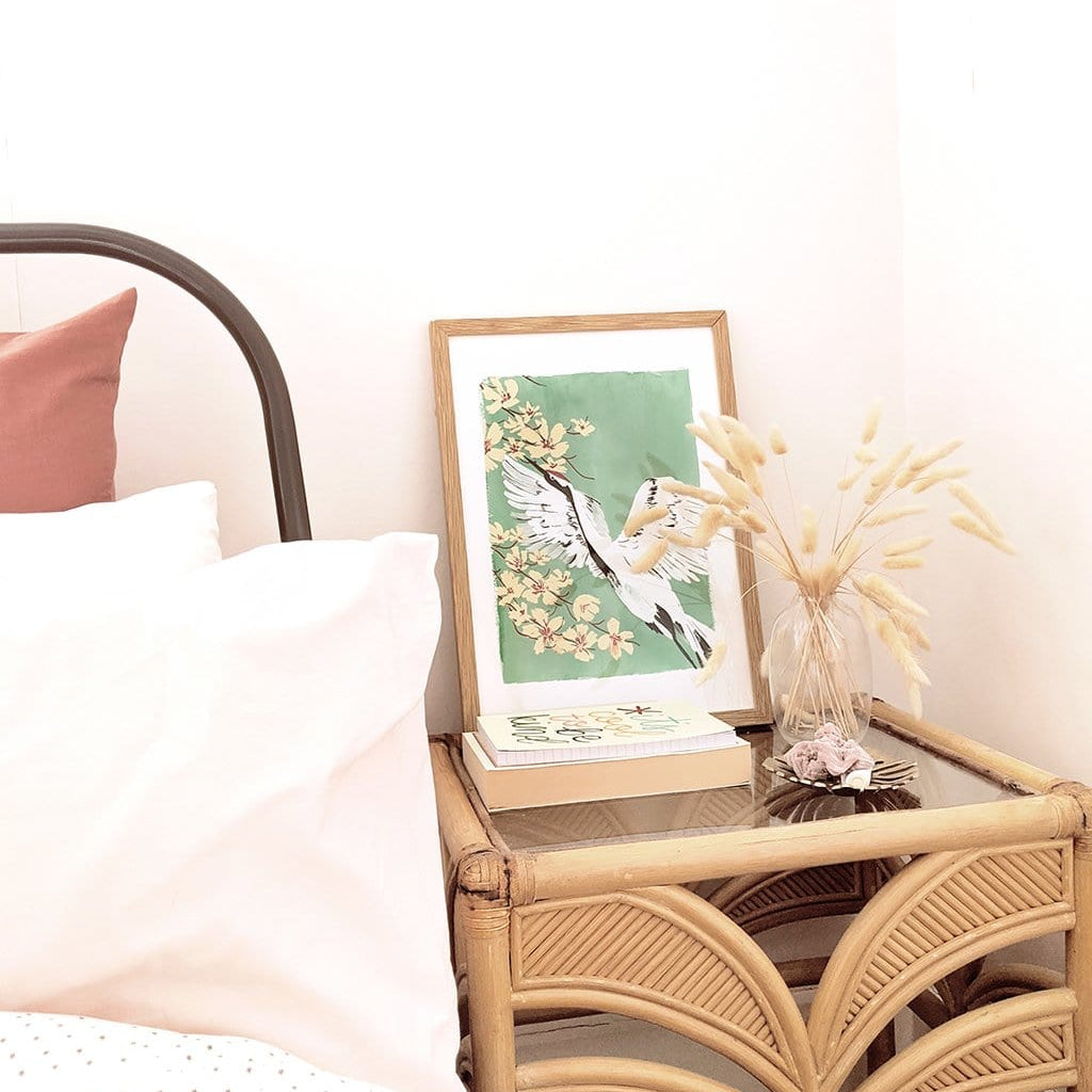 A Full Colour Giclee Print Featuring a Crane And Cherry Blossom On A Teal Background In A Light Oak Frame On A Side Table - Annie Dornan Smith