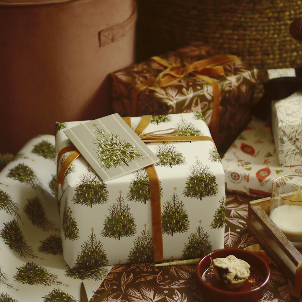 an illustrated christmas tree card rests on top of a present wrapped in matching wrapping paper, alongside lots of other beautiful gifts under the tree