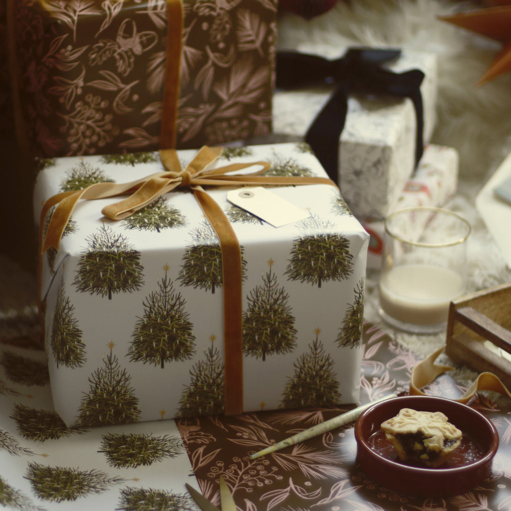 a gift wrapped with christmas tree patterned paper and tied with a gold velvet bow, alongside other beautiful gifts below the tree