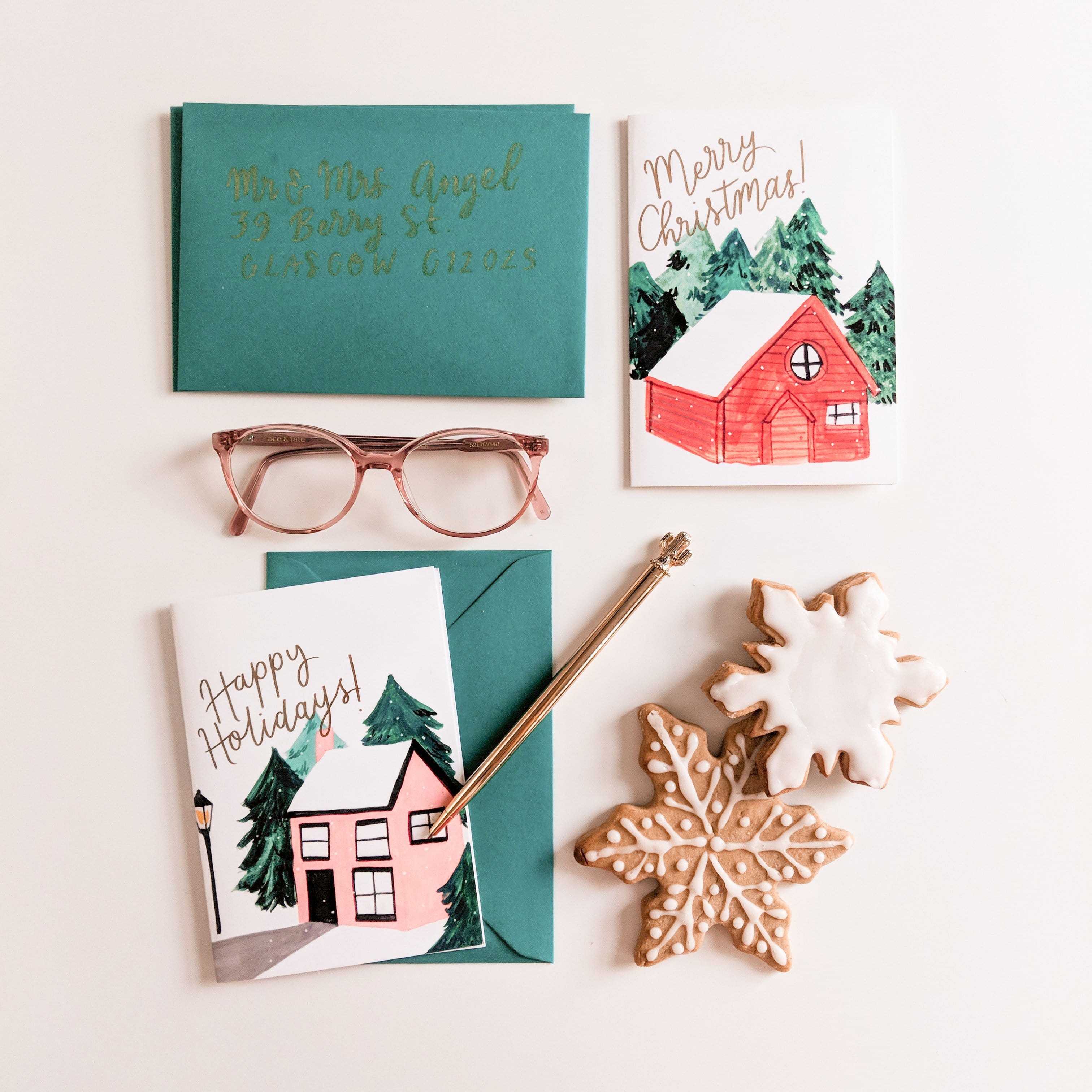 An Illustrated Snowy Cottage Christmas Card With Merry Christmas In Gold With A Teal Envelope With Christmas Biscuits - Annie Dornan Smith