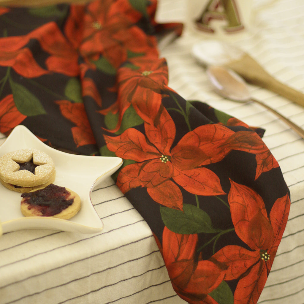 A cotton teatowel with a black and red poinsettia patterned print, on a christmassy table
