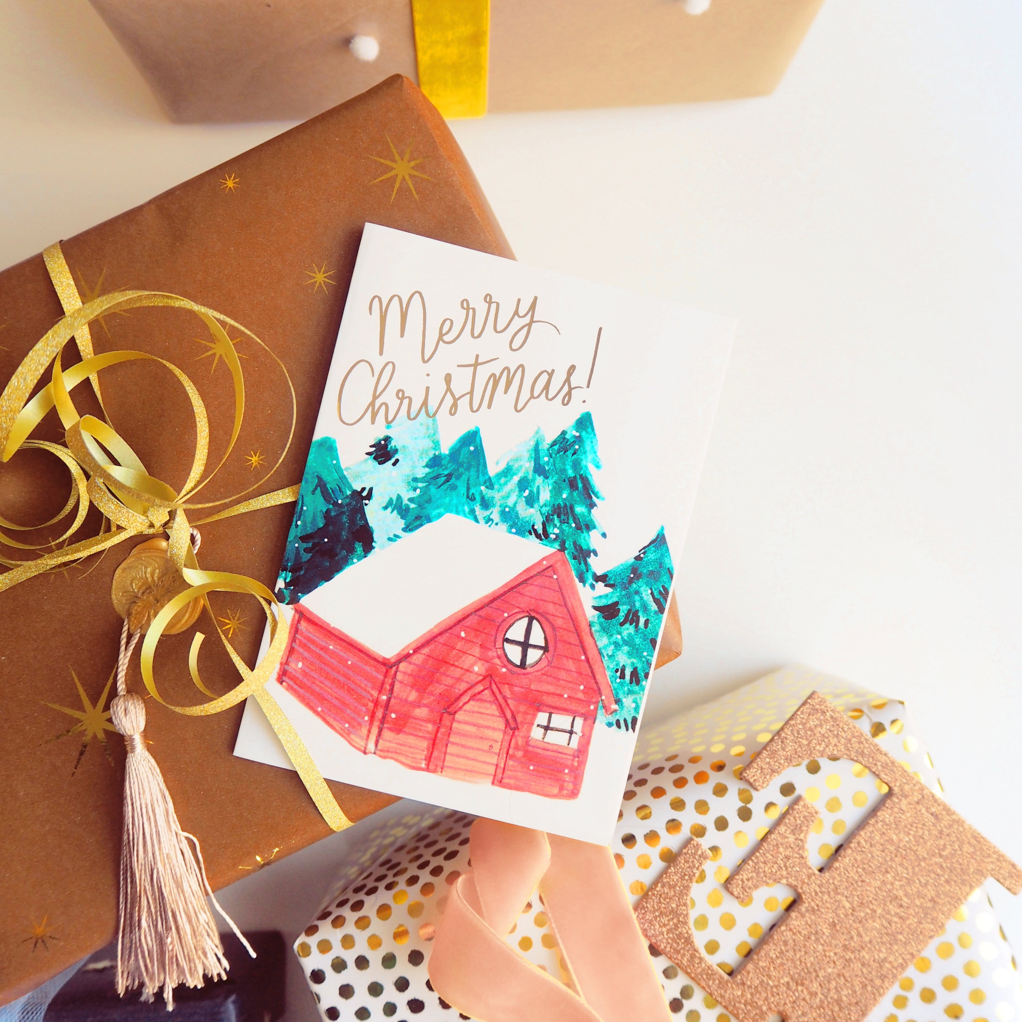 An Illustrated Snowy Chalet Christmas Card With Merry Christmas In Gold On Top Of A Christmas Gift - Annie Dornan Smith