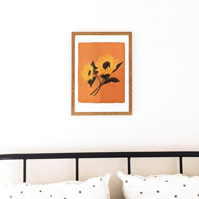 A Botanical Print Of Two Sunflowers On A Burnt Orange Background In A Light Oak Frame hung Above A Bed - Annie Dornan Smith