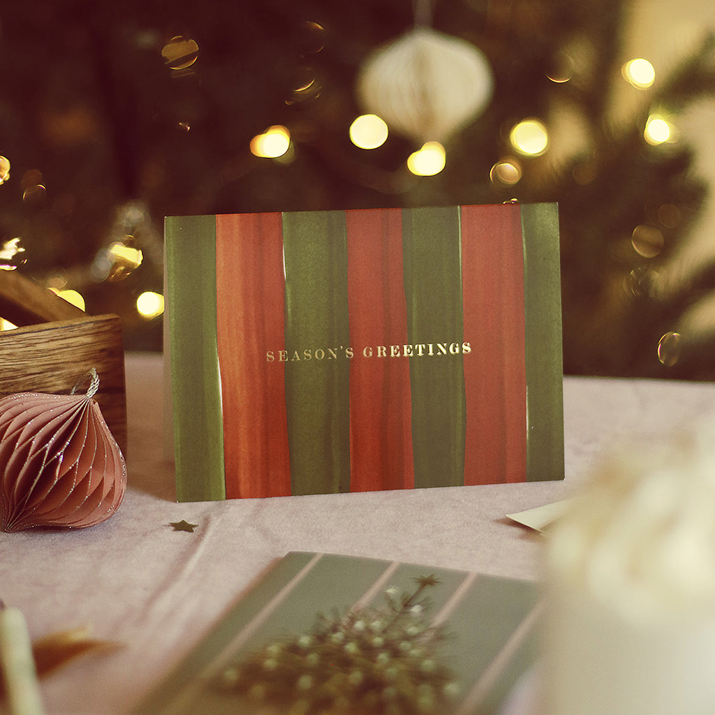 a red and green christmas card with handpainted stripes. Reads "seasons greetings" in gold foil