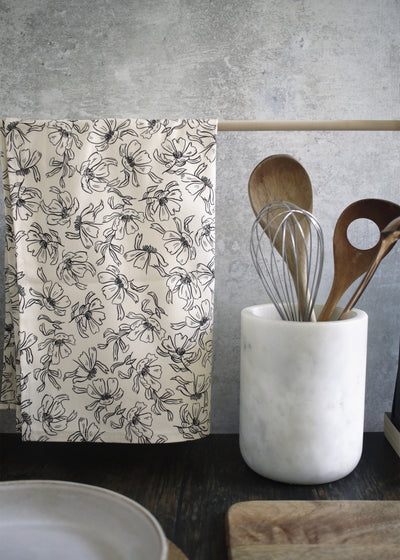 A black and cream floral patterned tea towel, hanging from a wooden rail on a countertop