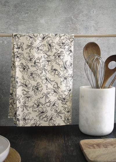 A black and cream floral patterned tea towel, hanging from a wooden rail on a countertop