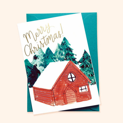 An Illustrated Snowy Chalet Christmas Card With Merry Christmas In Gold With A Teal Envelope - Annie Dornan Smith