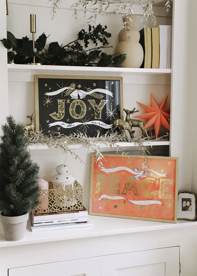Coral Christmas Print With Gold Peace Lettering And Stars With A White Ribbon And Dove In An Oak Frame With A Black Joy Print On A Book Shelf - Annie Dornan Smith