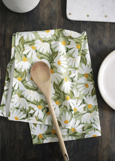a green cotton tea towel, covered in painted white daisy flowers, sitting on a dark wood table alongside plates and utensils