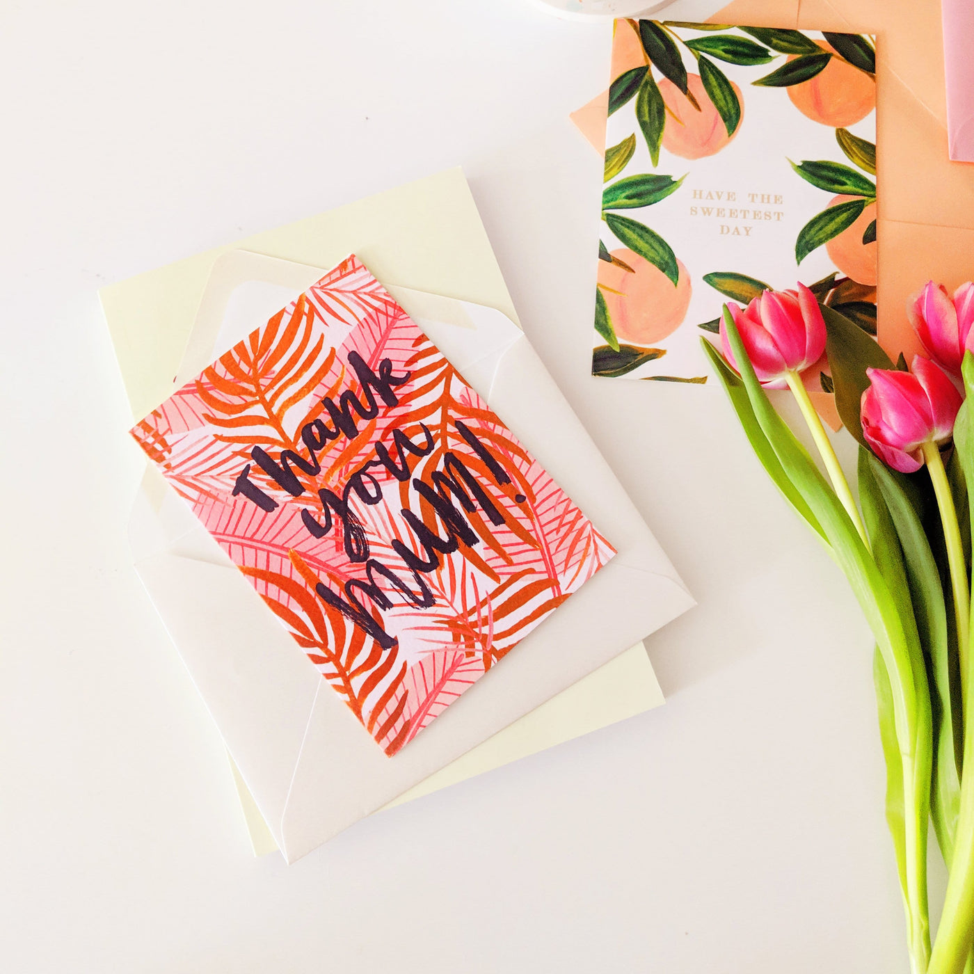 An Illustrated Pink Palm Leaf Card With The Words Thank You Mum In Brush Lettering With A White Envelope Next To Some Pink Tulips - Annie Dornan Smith