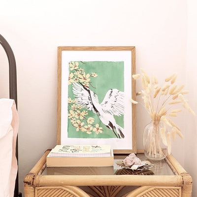 A Full Colour Giclee Print Featuring a Crane And Cherry Blossom On A Teal Background In A Light Oak Frame On A Side Table - Annie Dornan Smith