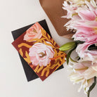 Botanical Greeting Card Pink English Tea Roses On Warm Brown Background With The Words From Dark Places Flowers Grow In Gold With Black Envelope Next To A Bunch Of Lilys - Annie Dornan Smith