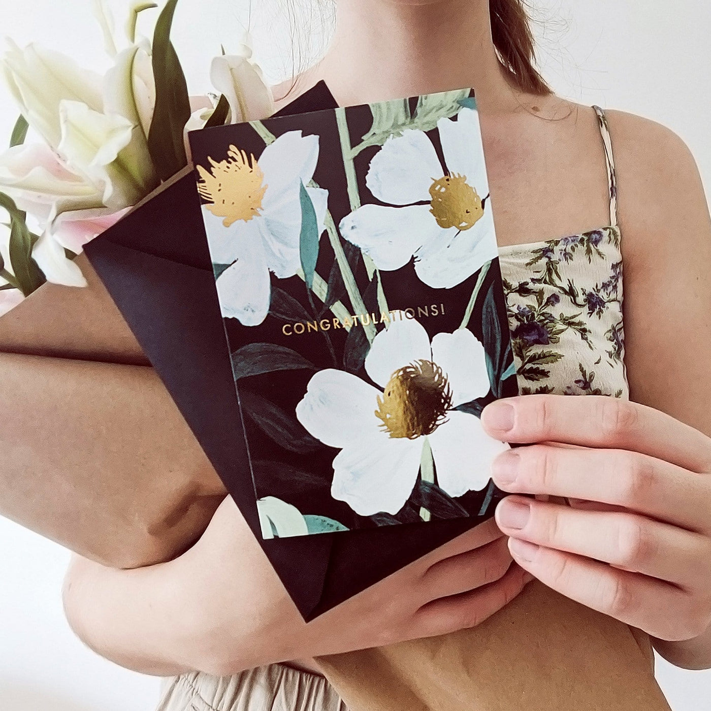 Girl Holding Illustrated White Flowers With Teal Leaves and Gold Centres On A Black Card With Congratulations In Gold With A Black Envelope - Annie Dornan Smith