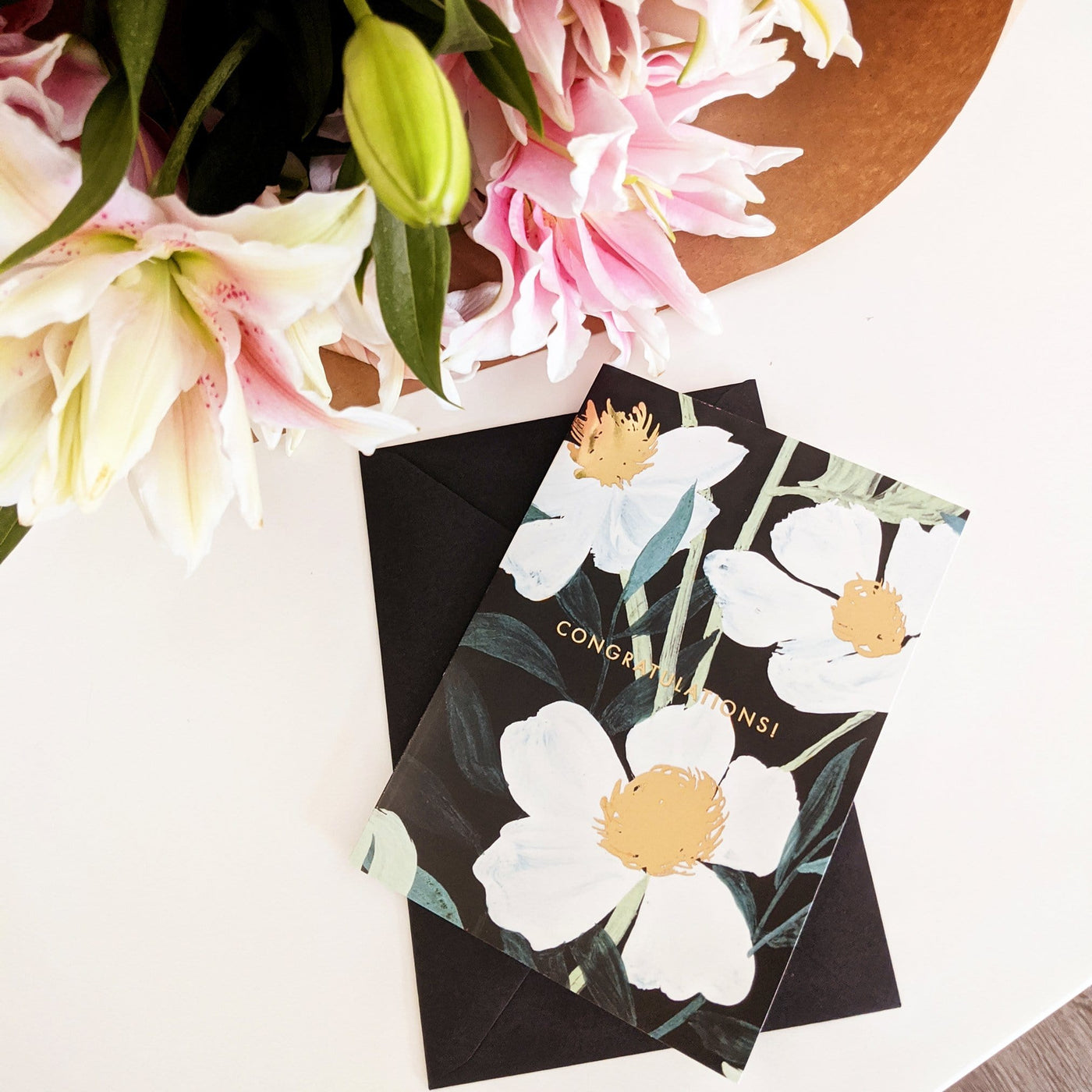 Illustrated White Flowers With Teal Leaves and Gold Centres On A Black Card With Congratulations In Gold With A Black Envelope On A White Table - Annie Dornan Smith