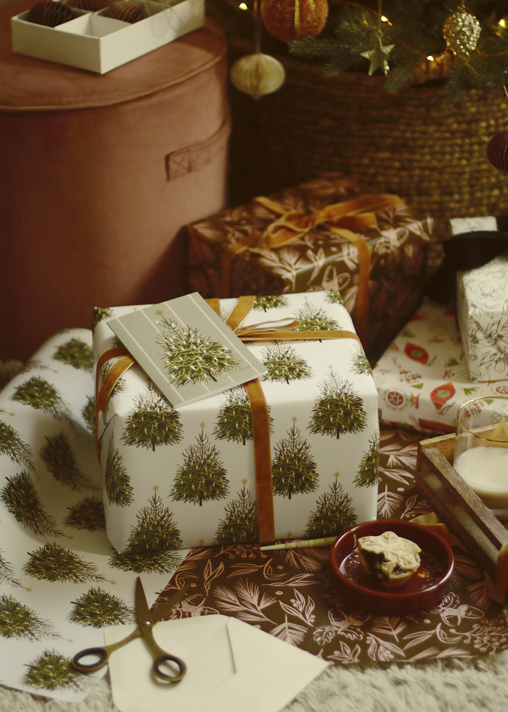  an illustrated christmas tree card rests on top of a present wrapped in matching wrapping paper, alongside lots of other beautiful gifts under the tree