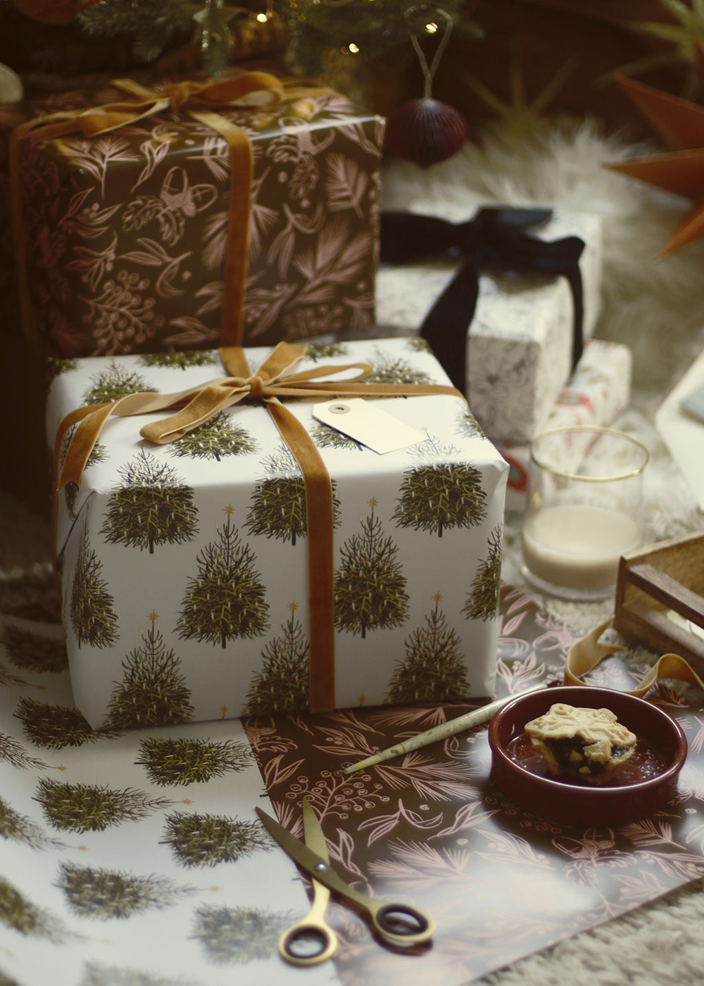 a gift wrapped with christmas tree patterned paper and tied with a gold velvet bow, alongside other beautiful gifts below the tree