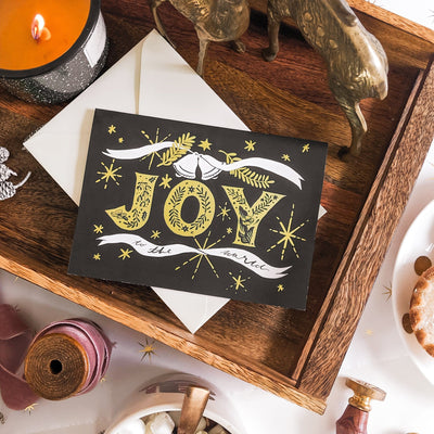 Black Christmas A6 Card With Gold Joy Lettering And Stars With Cream Envelope On A Dark Wood Tray - Annie Dornan Smith