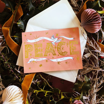 Coral Christmas Card With Gold Peace Lettering And Stars With A White Ribbon On A Bed Of Christmas Decorations - Annie Dornan Smith