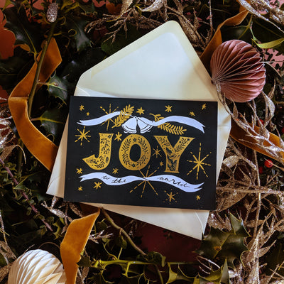 Black Christmas A6 Card With Gold Joy Lettering And Stars With Cream Envelope On A Pile Of Christmas Decorations - Annie Dornan Smith