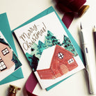 An Illustrated Snowy Chalet Christmas Card With Merry Christmas In Gold With A Teal Envelope - Annie Dornan Smith
