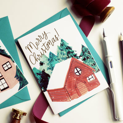 An Illustrated Snowy Chalet Christmas Card With Merry Christmas In Gold With A Teal Envelope - Annie Dornan Smith
