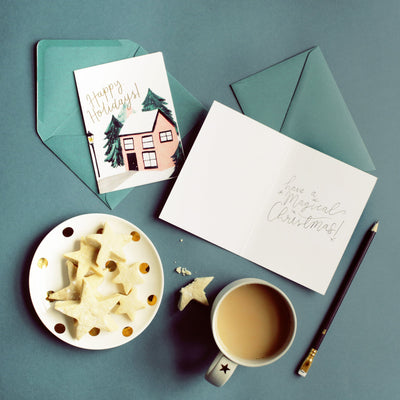 An Illustrated Snowy Cottage Christmas Card With Merry Christmas In Gold With A Teal Envelope With Star Biscuits And A Cup Of Tea - Annie Dornan Smith