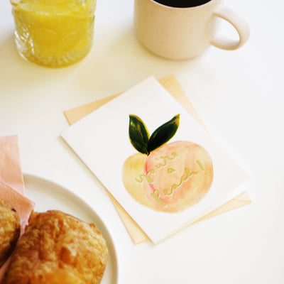 An Illustrated Care With A Peach And Green Leaf With The Words You're A Peach In Gold With A Peach Envelope Next To A Glass Of Orange Juice And Mug Of Coffee - Annie Dornan Smith