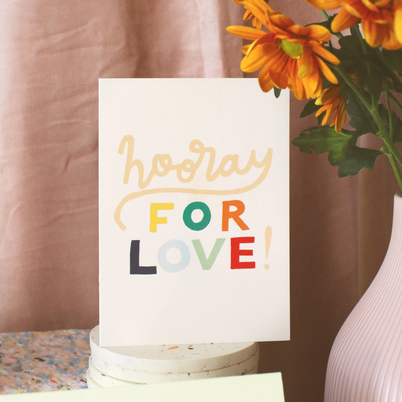 A Hand Lettered Rainbow Typography Card Which Reads Hooray For Love Next to Vase Of Orange Flowers - Annie Dornan Smith