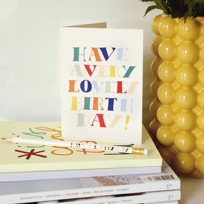 A Hand Lettered Rainbow Typography Card Reading Have A Very Lovely Birthday On A Pile Of Books - Annie Dornan Smith