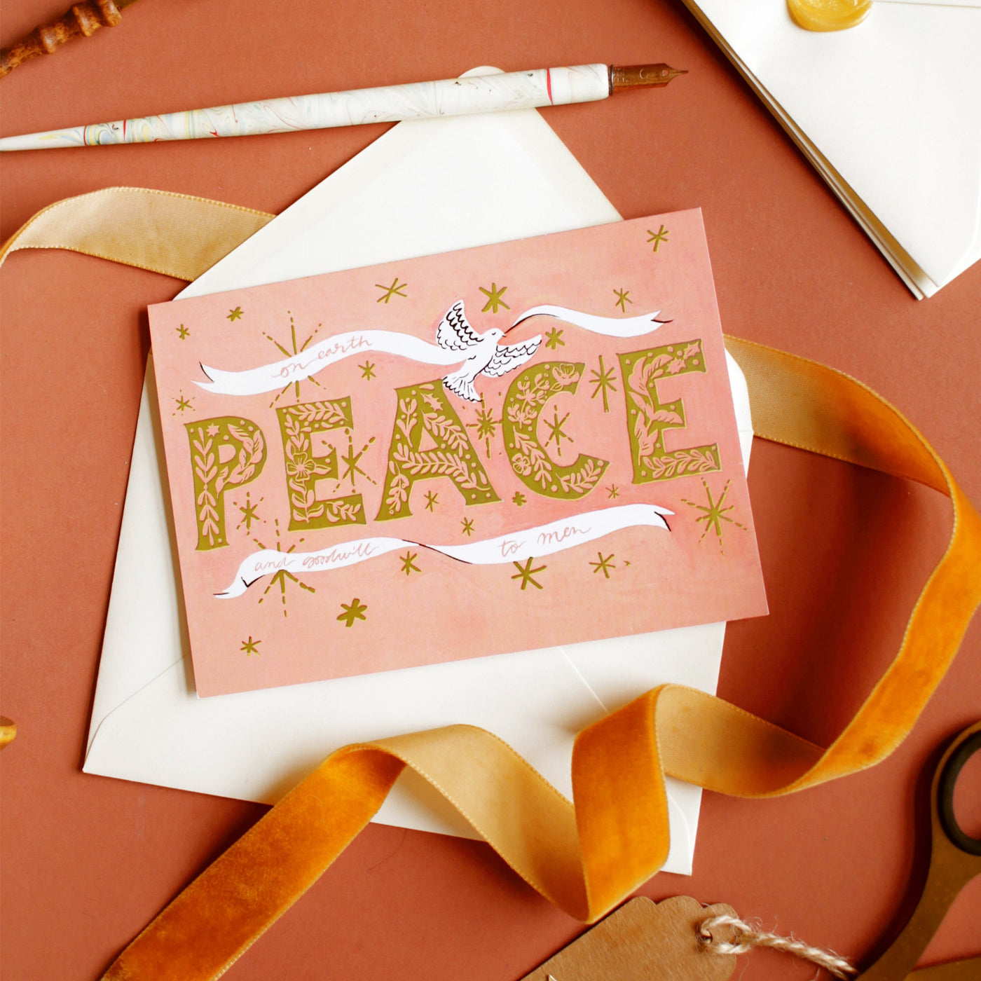 Coral Christmas Card With Gold Peace Lettering And Stars With A White Ribbon And Dove With A Gold Envelope With Orange Velvet Ribbon - Annie Dornan Smith
