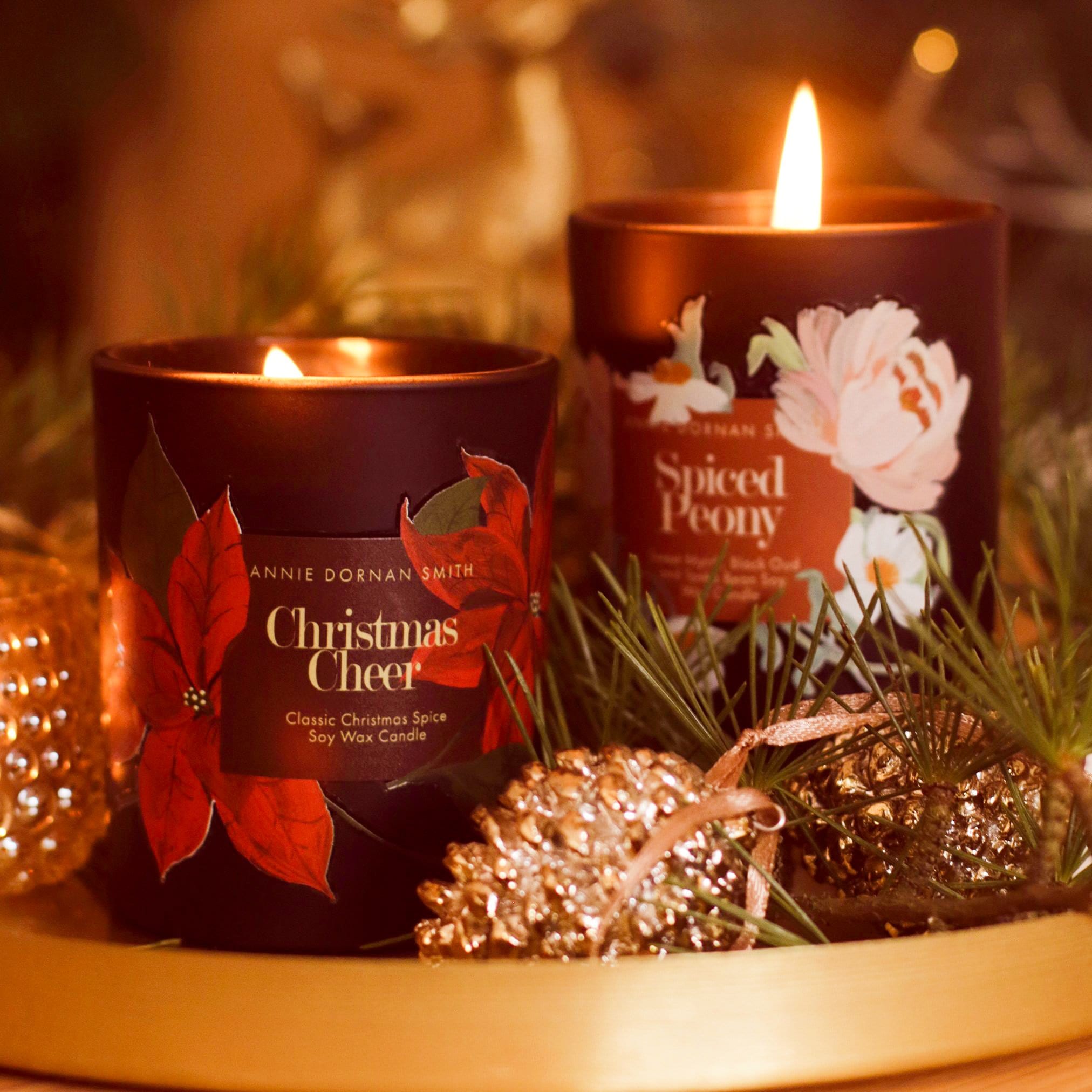 A Black Glass Candle Decorated with Red Poinsettias And A Christmas Cheer Label On A Gold Tray With Christmas Decorations  - Annie Dornan Smith