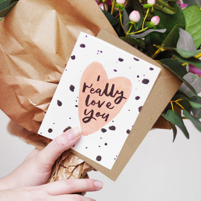 A Hand Lettered Card Which Reads I Really Love You On A Peach Heart With Black Paint Dots With A Kraft Envelope With A Bunch Of Flowers - Annie Dornan Smith