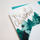 Close Up Of An Illustrated Snowy Chalet Christmas Card With Merry Christmas In Gold With A Teal Envelope - Annie Dornan Smith