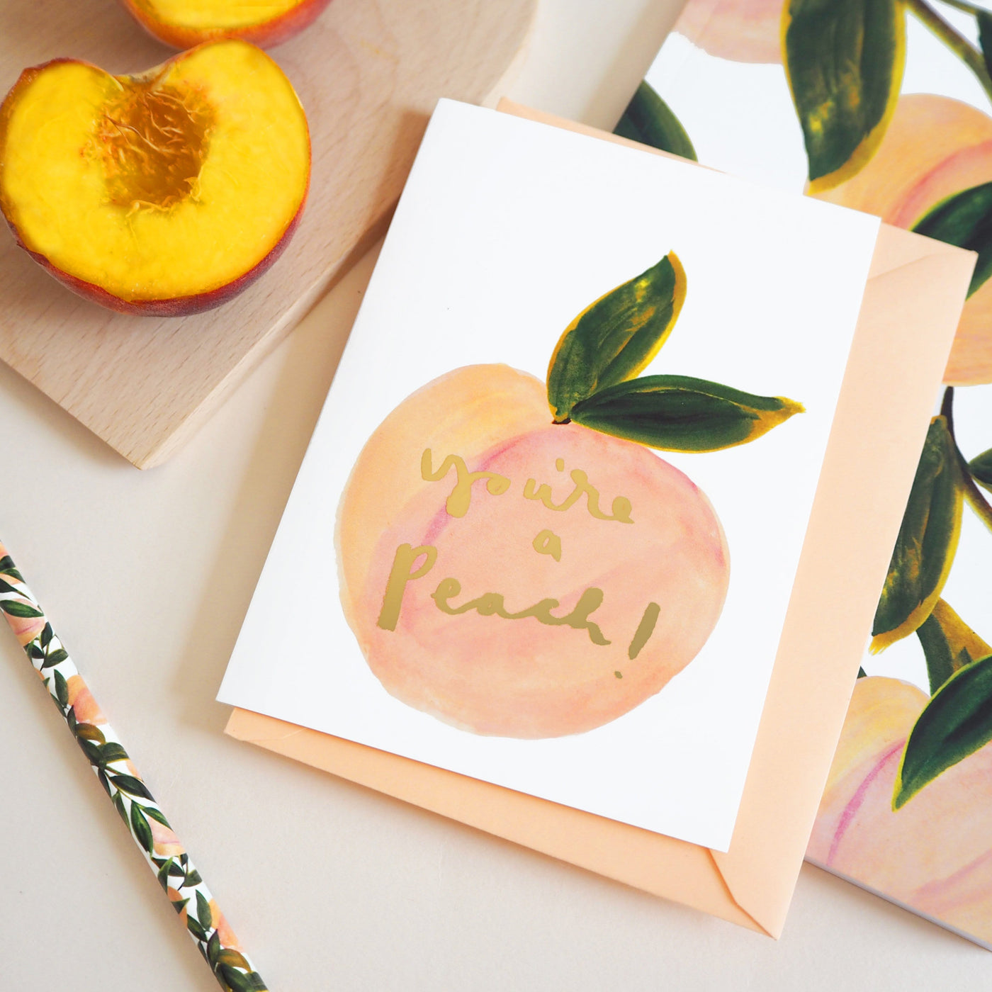 An Illustrated Care With A Peach And Green Leaf With The Words You're A Peach In Gold With A Peach Envelope Next To Real Peaches On A Wooden Board - Annie Dornan Smith