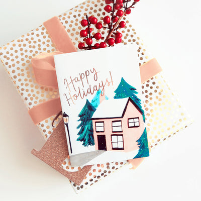 An Illustrated Snowy Cottage Christmas Card With Happy Holidays In Gold On Top Of A Wrapped Gift - Annie Dornan Smith