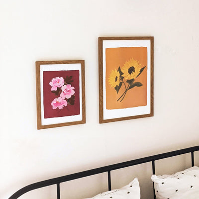 A Botanical Floral Print Of Three Pink Tea Roses On A Deep Pink Background In An Oak Frame Hanging On A Wall Next To A Sunflower Print - Annie Dornan Smith