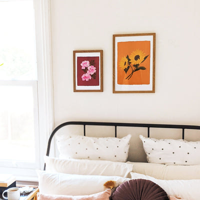 A Botanical Print Of Two Sunflowers On A Burnt Orange Background In A Light Oak Frame Hung Above A Bed Next To Another Spring Florals Print - Annie Dornan Smith