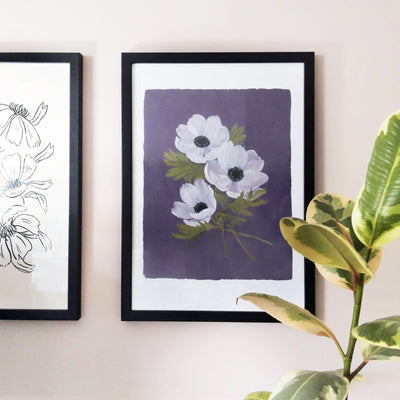A purple floral print in a black frame hanging on a pale pink wall, alongside a rubber plant