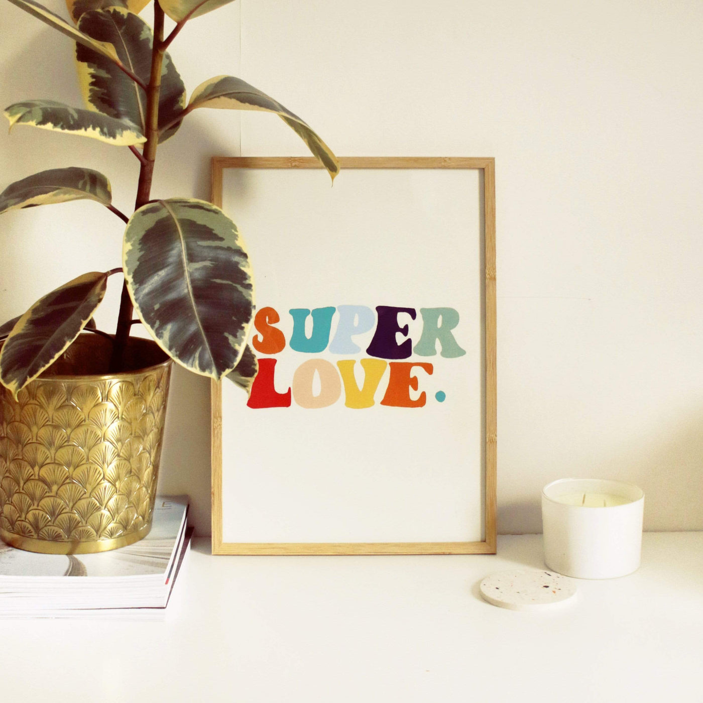 A Hand Lettered Rainbow Typography Print Which Reads Super Love In A Light Oak Frame Next To A Plant - Annie Dornan Smith