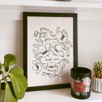 a minimal, line art flower print with quote "forever and ever" in a black frame on a bookshelf, alongside a candle and houseplants